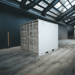 White shipping container in a large industrial warehouse with steel beams and skylights above.