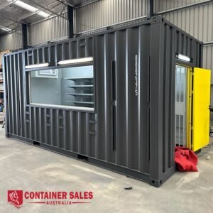 Grey modular shipping container with a large window and interior shelves, yellow door at the far end, inside a warehouse.