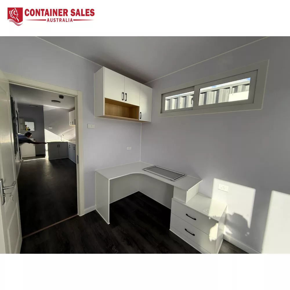 40ft High Cube Accommodation Container Home 5 Bright home office nook with an L-shaped white desk, wall cabinets, and a window above; visible doorway to a kitchen in the background.