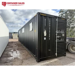 Black shipping container in a gravel yard with serial numbers on the door, under a cloudy sky.