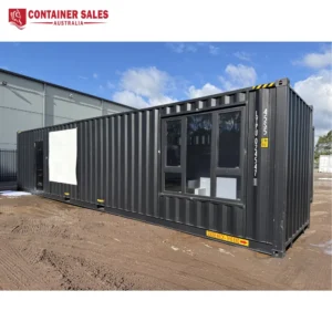 Dark gray shipping container converted into an office with large glass windows and a door on a dirt lot under a blue sky.