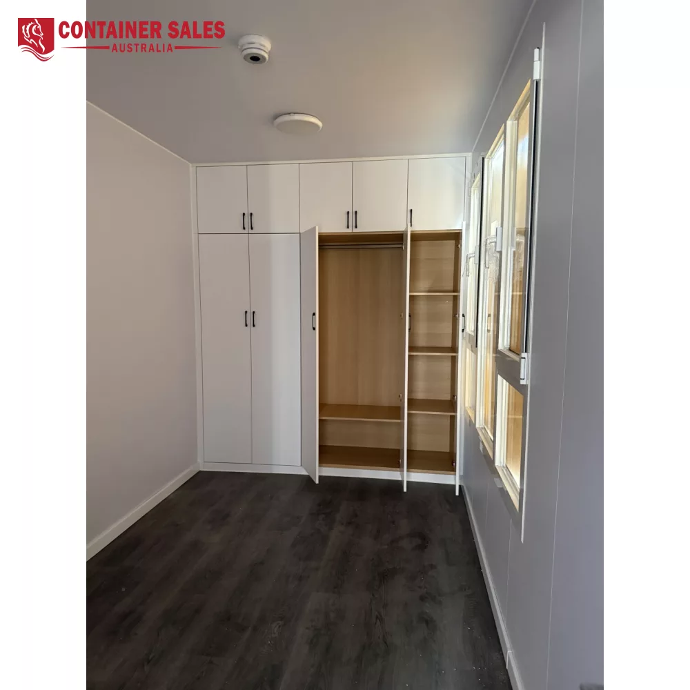 40ft High Cube Accommodation Container Home 4 White built-in wardrobes with tall cupboards and an open section showing wooden shelves in a small, grey-walled room with dark wood flooring and windows on the right.