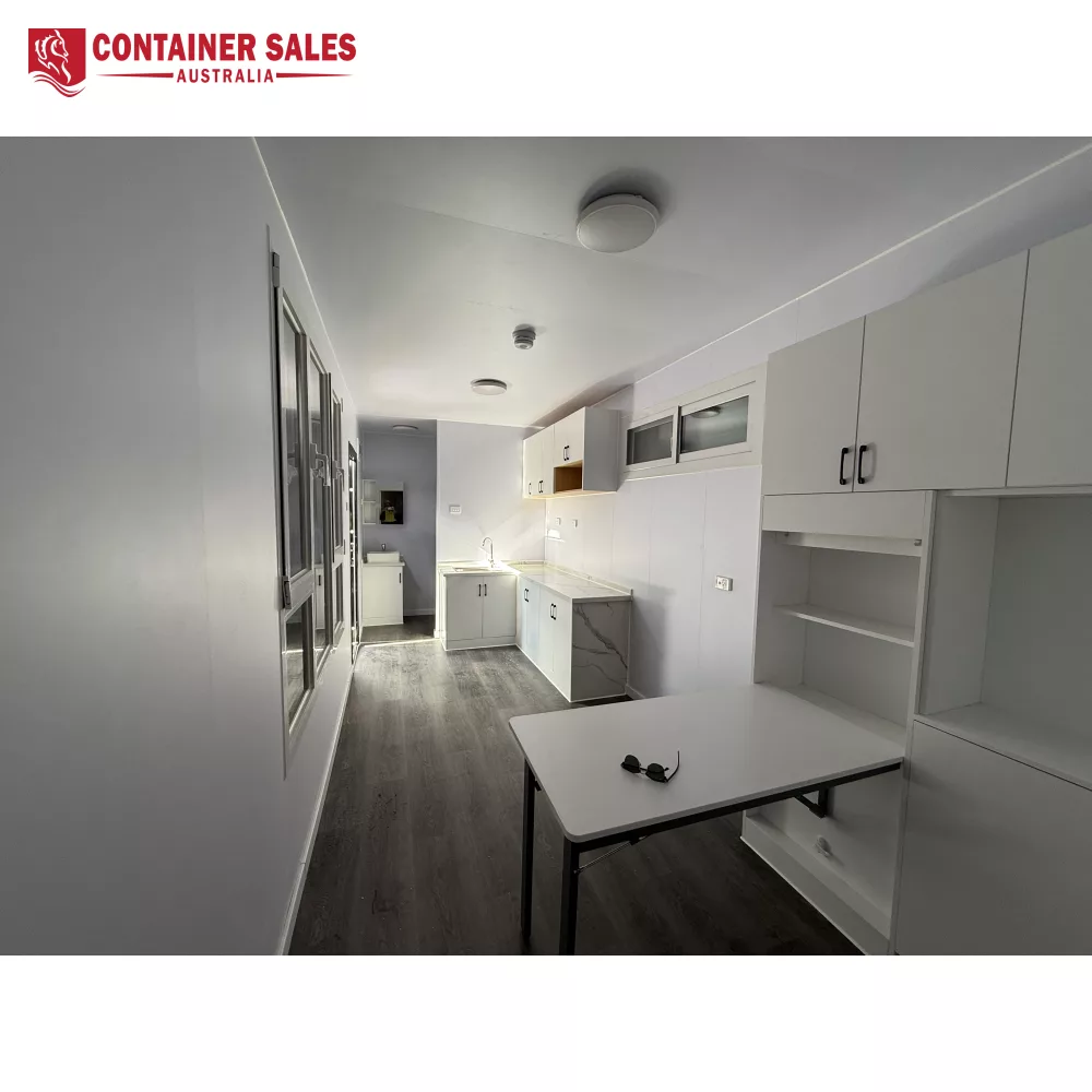 40ft High Cube Accommodation Container Home 3 Bright, minimalist kitchen with white cabinets, grey countertops, and a small white table with sunglasses on top in the foreground.