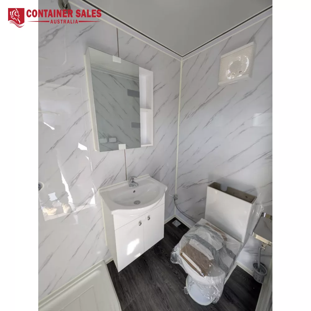 4m Accommodation Unit (Box) – Portable Cabin 3 Small modern bathroom with white vanity and mirror cabinet, marble walls, and a plastic-wrapped toilet; a wall vent is visible near the ceiling.