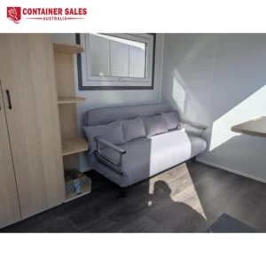 Small living area inside a compact container home: gray sofa bed with cushions beside a wooden cabinet and shelves under a window.