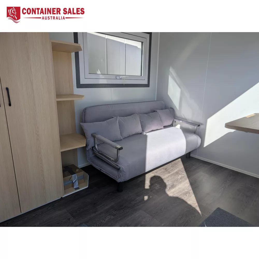 4m Accommodation Unit (Box) – Portable Cabin 2 Small living area inside a compact container home: gray sofa bed with cushions beside a wooden cabinet and shelves under a window.