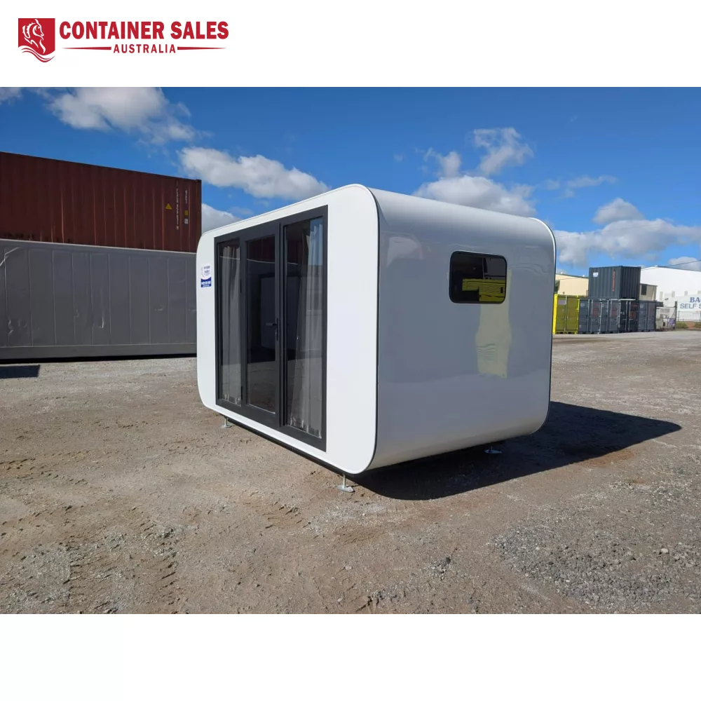4m Pod House – Backyard Studio Pod 1 White rounded modular container with large glass doors on a gravel lot under a blue sky; shipping containers in the background.