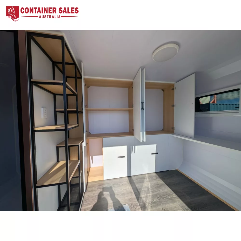 4m Pod House – Backyard Studio Pod 4 Interior of a storage trailer showing open upper cabinets with light wood shelves and lower white drawers; a black metal shelving unit sits along the left wall; a window on the right brings in light, with grey flooring visible.