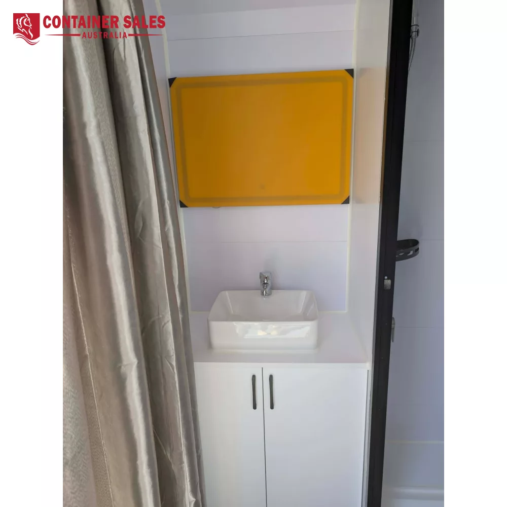 4m Pod House – Backyard Studio Pod 3 White bathroom vanity with a square white sink and chrome faucet, orange-framed mirror panel above, gray curtains to the left, and a dark door frame to the right.