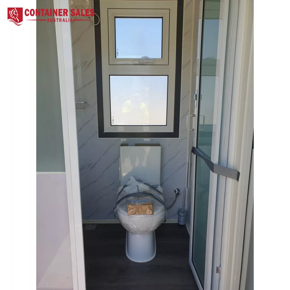 6m 2-Room Accommodation Unit (Type C) 4 Toilet in a small, waterproof bathroom under installation, wrapped in plastic with a cardboard cover on the seat, next to a window and glass door.