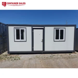 Front view of a grey modular container with two white-framed windows and a central door on a gravel lot.