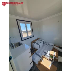 Interior of a small prefab container home: kitchenette with sink on the left, a bed frame wrapped in packaging on the right, window viewing outdoors.