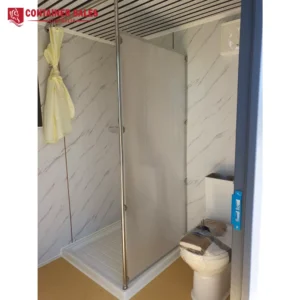 Compact bathroom inside a shipping container: corner shower with frosted glass door, marble-pattern walls, and a wrapped toilet.