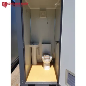 Portable restroom inside a compact metal cabin: pedestal sink on the left and a wrapped toilet on the right, with a vent on the back wall above.